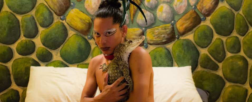 A person holding an alligator to their chest, their chest bare, and they’re looking straight into the camera with serious eyes.